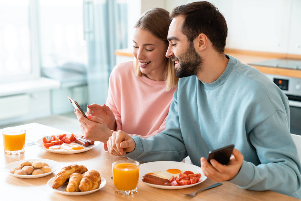 自宅で居心地の良いキッチンで朝食をしながら、携帯電話や笑顔を使用して若い喜びのカップルの肖像画 - 写真・画像