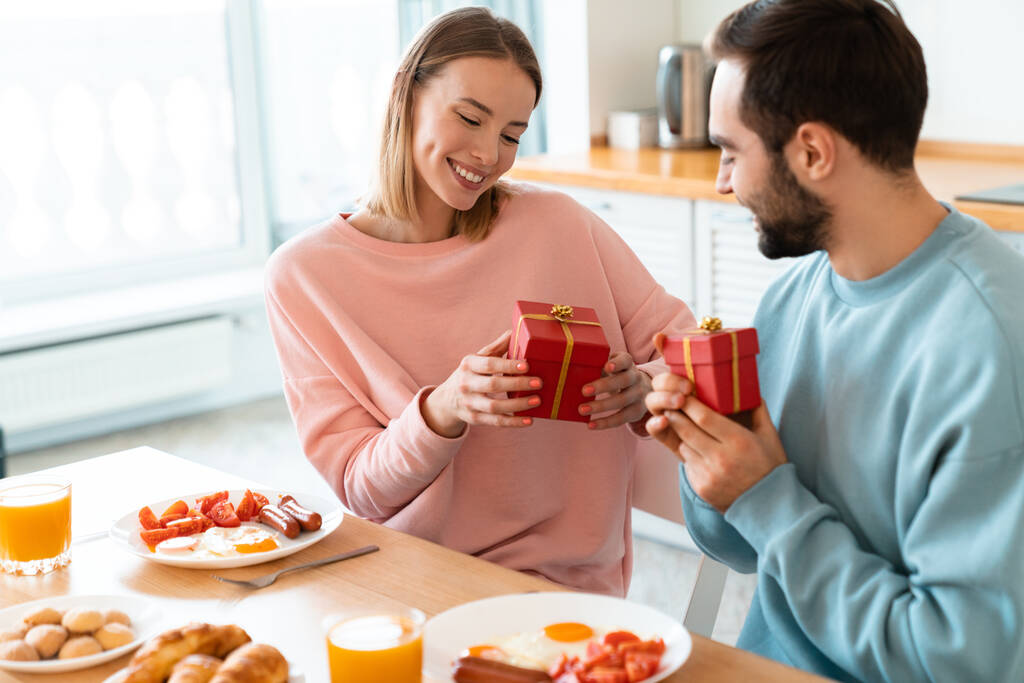 Porträt eines glücklichen jungen Paares mit Geschenkschachteln beim Frühstück in der gemütlichen Küche zu Hause - Foto, Bild