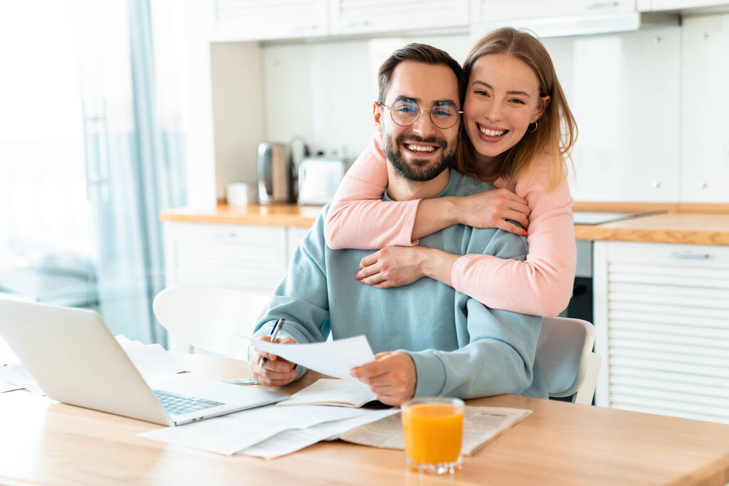 Portrait de jeune couple souriant étreignant tout en travaillant avec un ordinateur portable et des documents dans une cuisine confortable à la maison
 - Photo, image