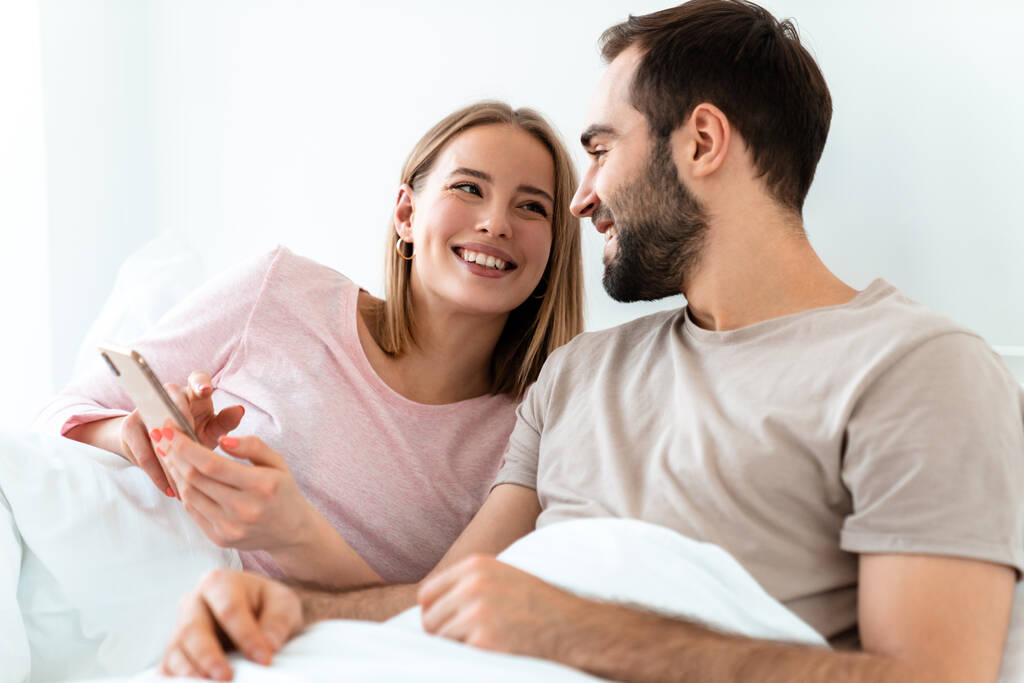 美しい喜びのカップルの笑顔と自宅の寝室に横たわっている間、スマートフォンを使用しての肖像画 - 写真・画像