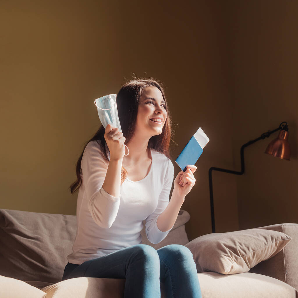 cheerful young woman holding passport with air ticket and medical mask while sitting on sofa, end of quarantine concept - Photo, Image