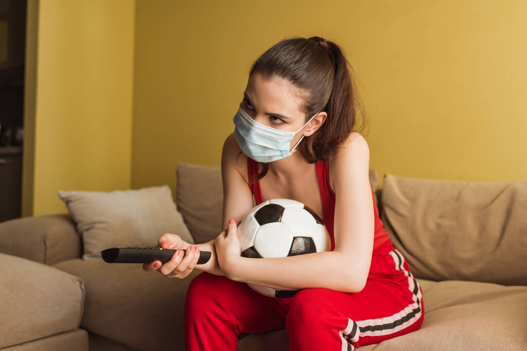 Sportliches Mädchen mit medizinischer Maske hält Fernbedienung in der Nähe des Fußballs, während sie die Meisterschaft im Wohnzimmer verfolgt  - Foto, Bild