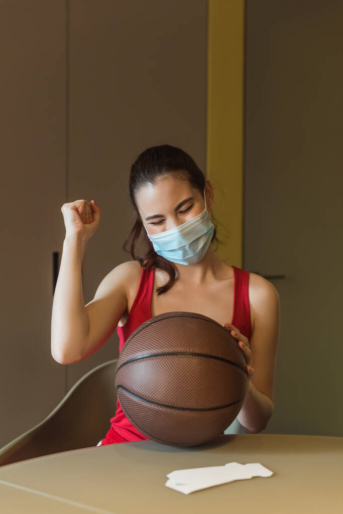 sportieve vrouw met medisch masker en gesloten ogen juichen in de buurt van basketbal - Foto, afbeelding