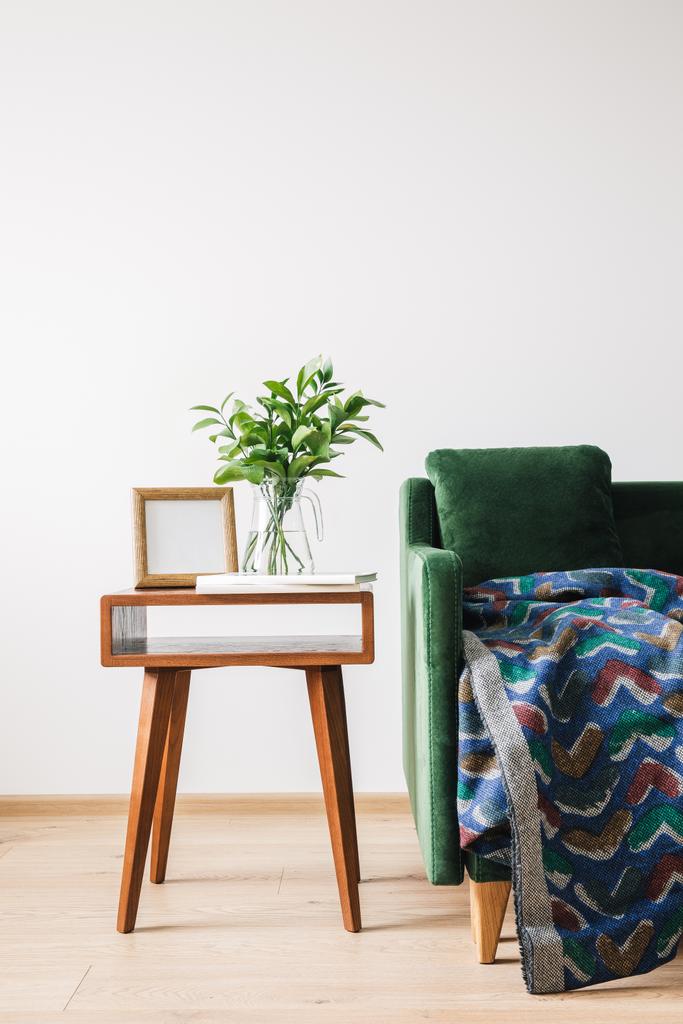 grünes Sofa mit Kissen und Decke neben Holztisch mit grüner Pflanze, Büchern und Fotorahmen - Foto, Bild