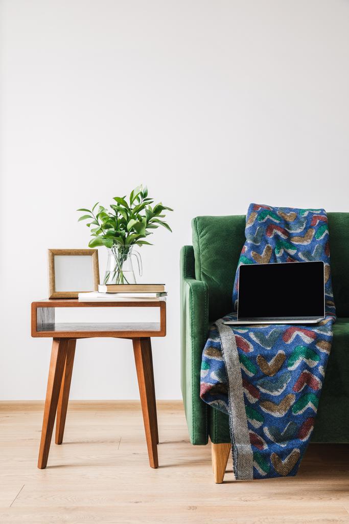 zöld kanapé takaróval és laptoppal fa dohányzóasztal mellett, zöld növényekkel, könyvekkel és képkerettel - Fotó, kép