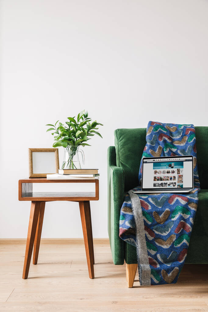 KYIV, UKRAINE - APRIL 14, 2020: green sofa with blanket and laptop with amazon website near wooden coffee table with green plant, books and photo frame - Photo, Image