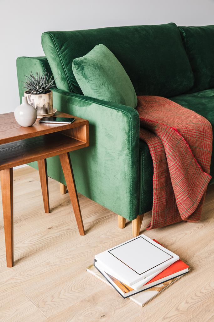 зеленый диван с одеялом рядом с деревянным журнальным столиком с растением и смартфоном рядом с книгами на полу
 - Фото, изображение