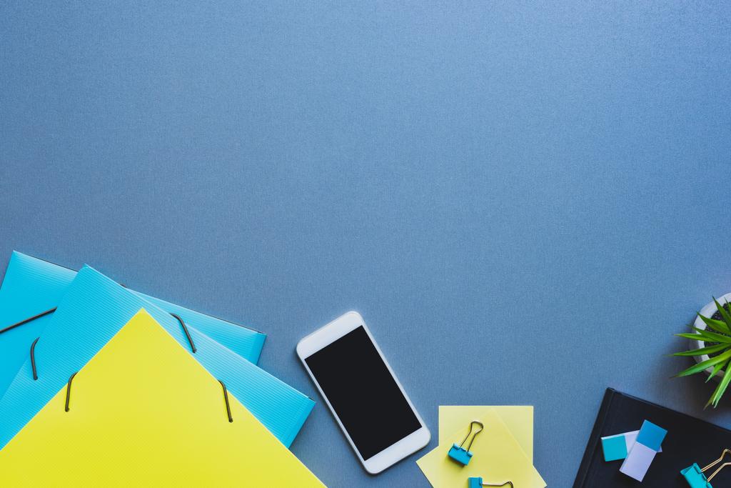 Вид сверху смартфона с бланковым экраном рядом с бумажными папками и канцелярскими принадлежностями на синем фоне
 - Фото, изображение