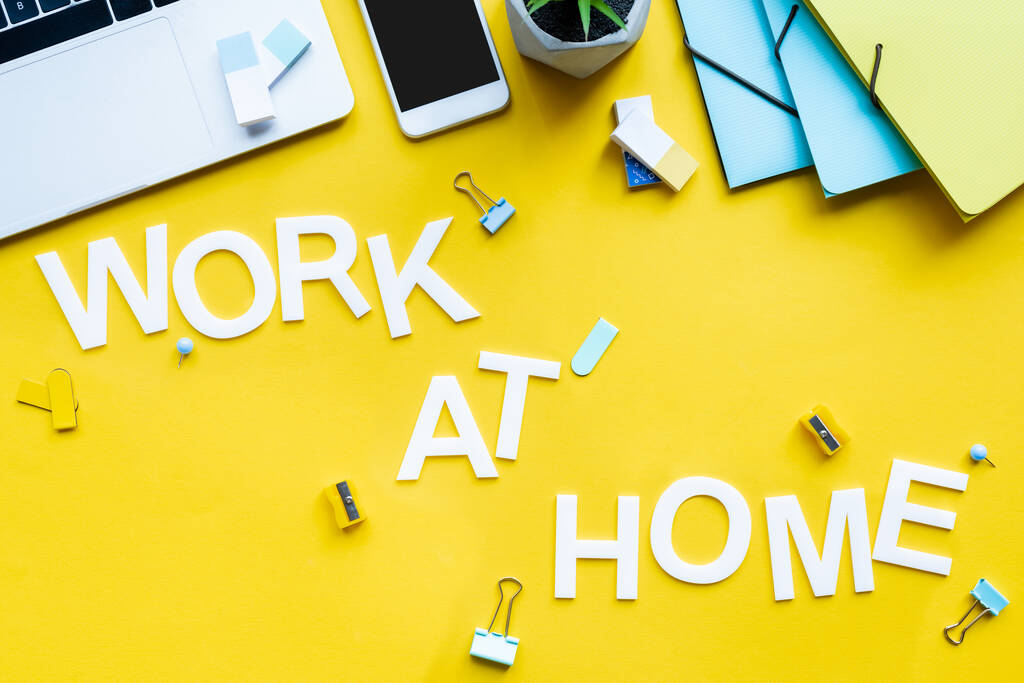 Vista superior do trabalho em casa lettering perto de smartphones, laptop e material de escritório na superfície amarela
 - Foto, Imagem