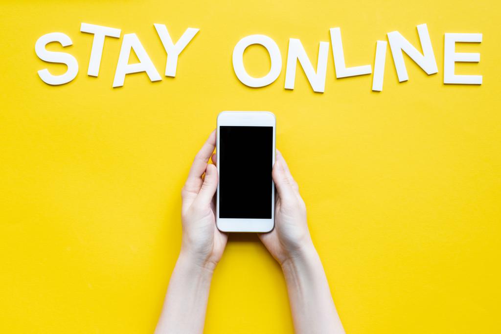Vista recortada de la mujer sosteniendo el teléfono inteligente con pantalla en blanco y letras permanecer en línea en la superficie amarilla
 - Foto, Imagen