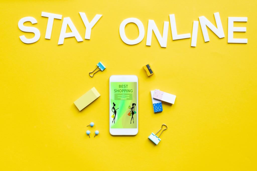 Visão superior do smartphone com o melhor aplicativo de compras perto de materiais de escritório e ficar lettering on-line na superfície amarela
 - Foto, Imagem