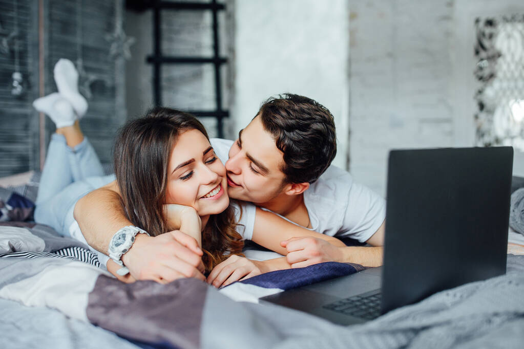 若い結婚し、素敵なカップルは自宅でベッドの上に横たわって、ラップトップを使用しています。完璧な朝だ! - 写真・画像