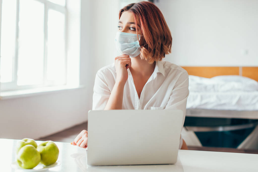 female pensive freelancer in medical mask working on laptop at home with apples on quarantine - Photo, Image