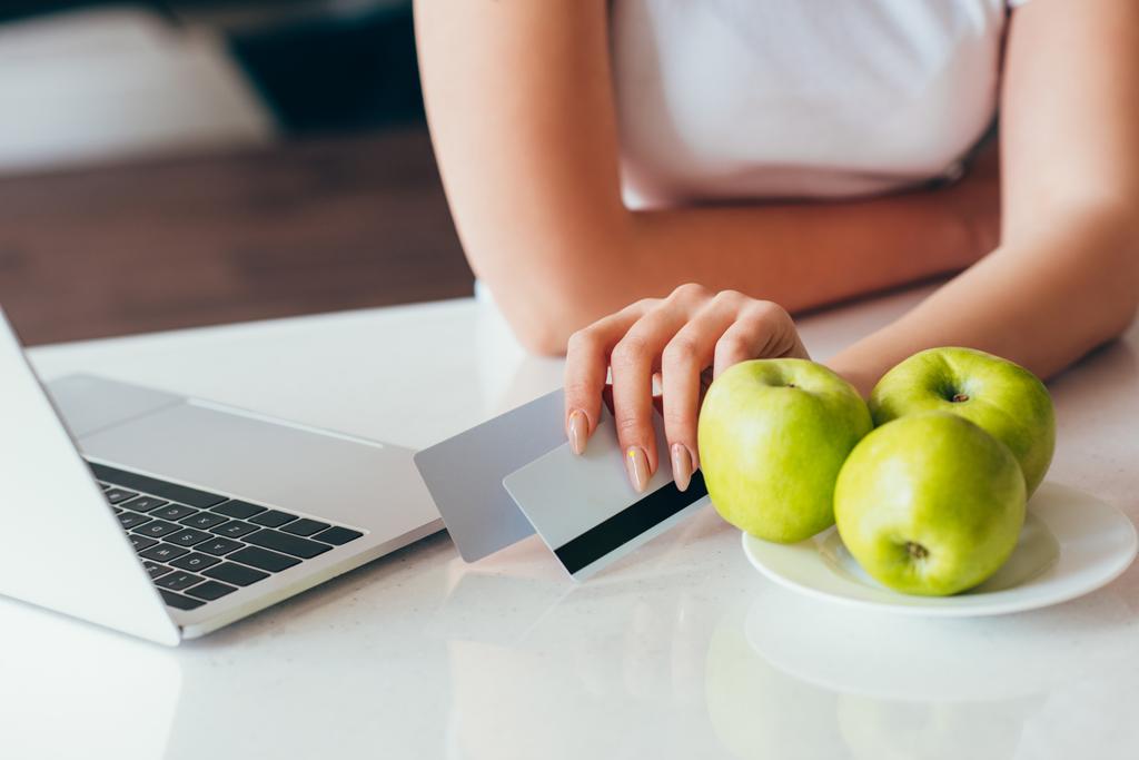 przycięty widok kobiety zakupy online z laptopem i kartami kredytowymi na kuchni z jabłkami podczas samodzielnej izolacji  - Zdjęcie, obraz