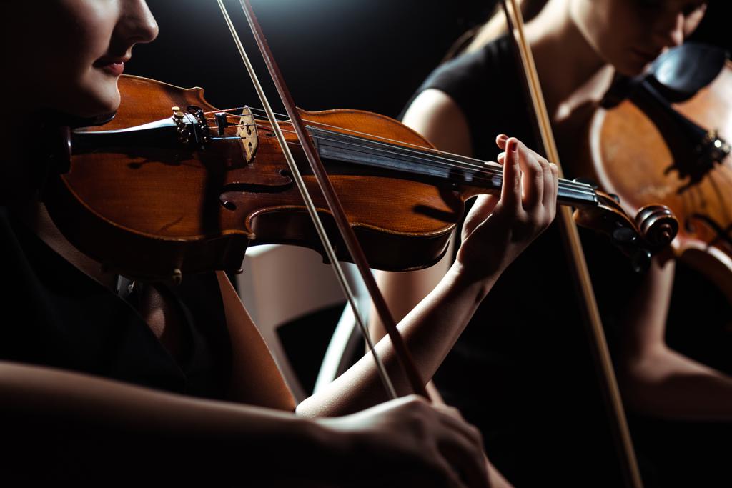 bijgesneden mening van twee vrouwelijke professionele muzikanten spelen op violen op donker podium  - Foto, afbeelding