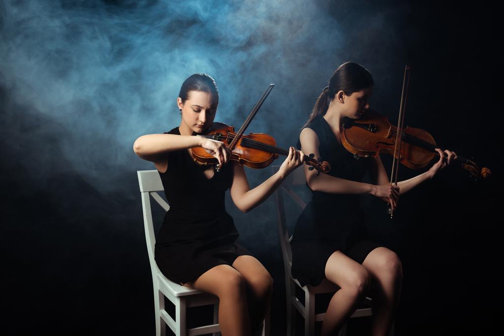 attractive female musicians playing on violins on dark stage with smoke - Photo, Image