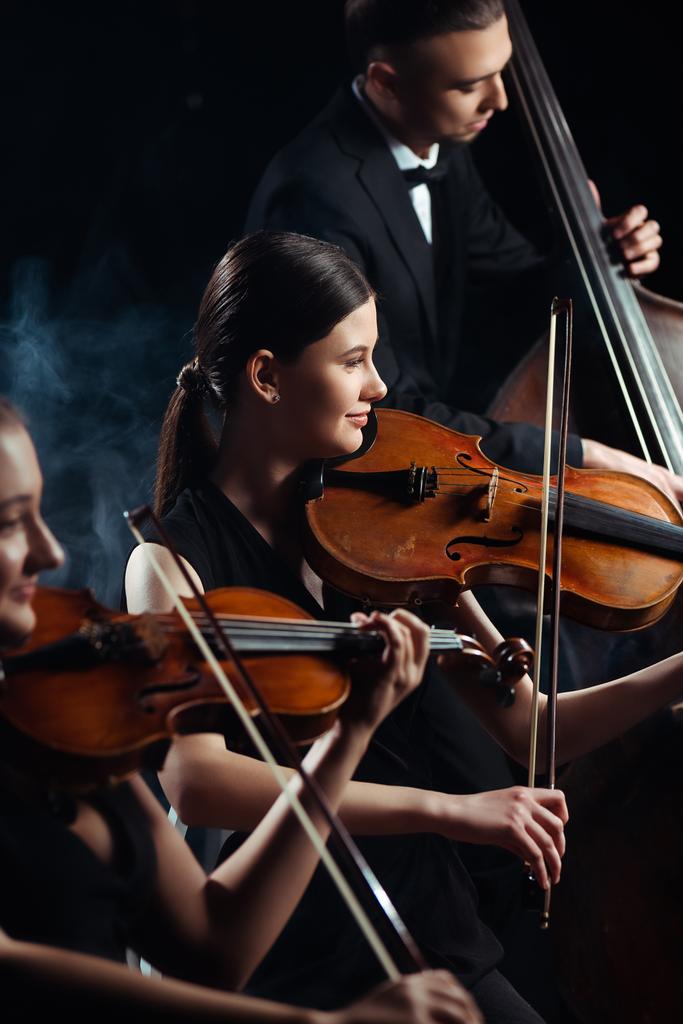 Trio professioneller Musiker spielt auf Geigen und Kontrabass auf dunkler Bühne mit Rauch - Foto, Bild