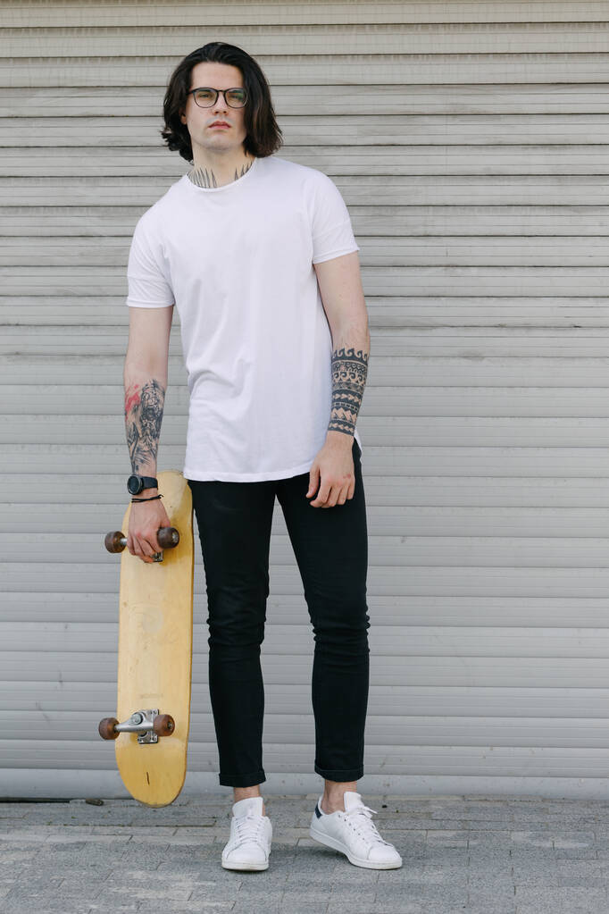 Hipster bel modello maschile indossa una t-shirt bianca bianca con spazio per il tuo logo o design in stile urbano casual. Sta tenendo uno skateboard.
 - Foto, immagini