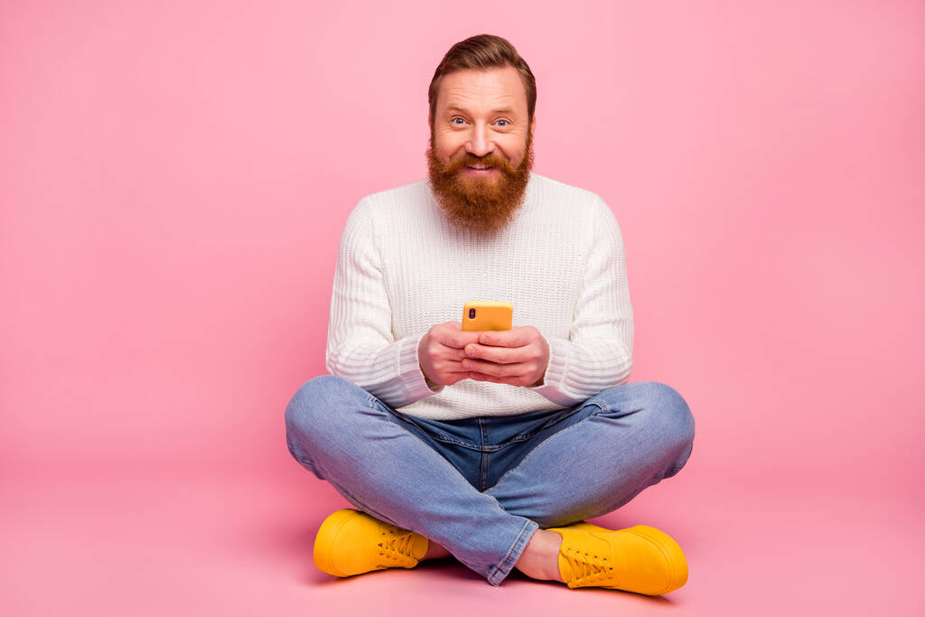 Фото позитивного веселого чоловіка сидять на підлозі ноги схрещені за допомогою мобільного телефону читати новини в соціальних мережах смс-повідомлення, надягати джинси-джампінг модне взуття ізольоване пастельний кольоровий фон
 - Фото, зображення