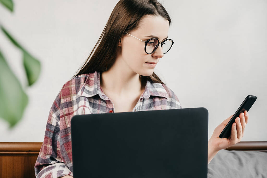 Widok z przodu młodej kobiety w okularach za pomocą aplikacji biznesowych na smartfonie z laptopem siedzącym na wygodnym łóżku w domu, kobiece ręce trzymające telefon komórkowy sprawdzające wiadomości online - Zdjęcie, obraz