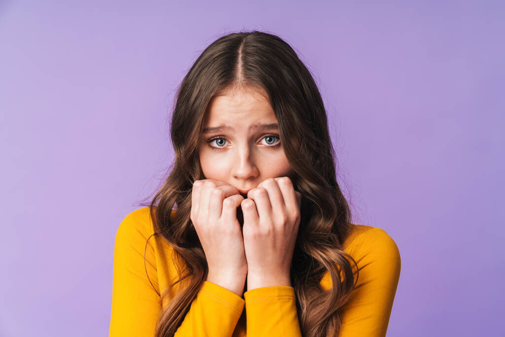 Imagem de uma jovem caucasiana com cabelo castanho comprido a ter medo e a morder as unhas isoladas sobre fundo violeta
 - Foto, Imagem