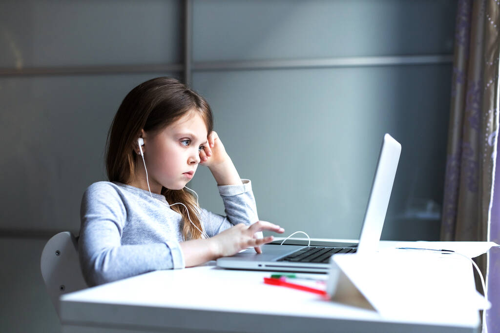 Zmęczony internetowymi zajęciami komputerowymi noszącymi słuchawki, dziecko patrzy na monitor ze smutkiem. Dziewczyna z realizacji pracy domowej. Uczennica w domu z laptopem. - Zdjęcie, obraz