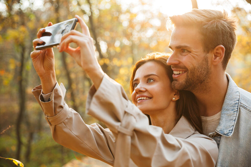 Bella giovane coppia innamorata usando il cellulare mentre trascorre del tempo al parco autunnale, scattando un selfie
 - Foto, immagini