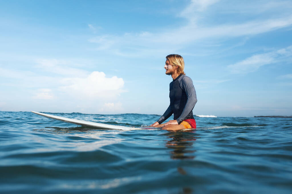 Surfeando. Hombre Surfista Sentado en Retrato de Tabla de Surf. Guapo con traje de neopreno en el océano. Mar azul y hermoso cielo con nubes suaves en el fondo
. - Foto, Imagen