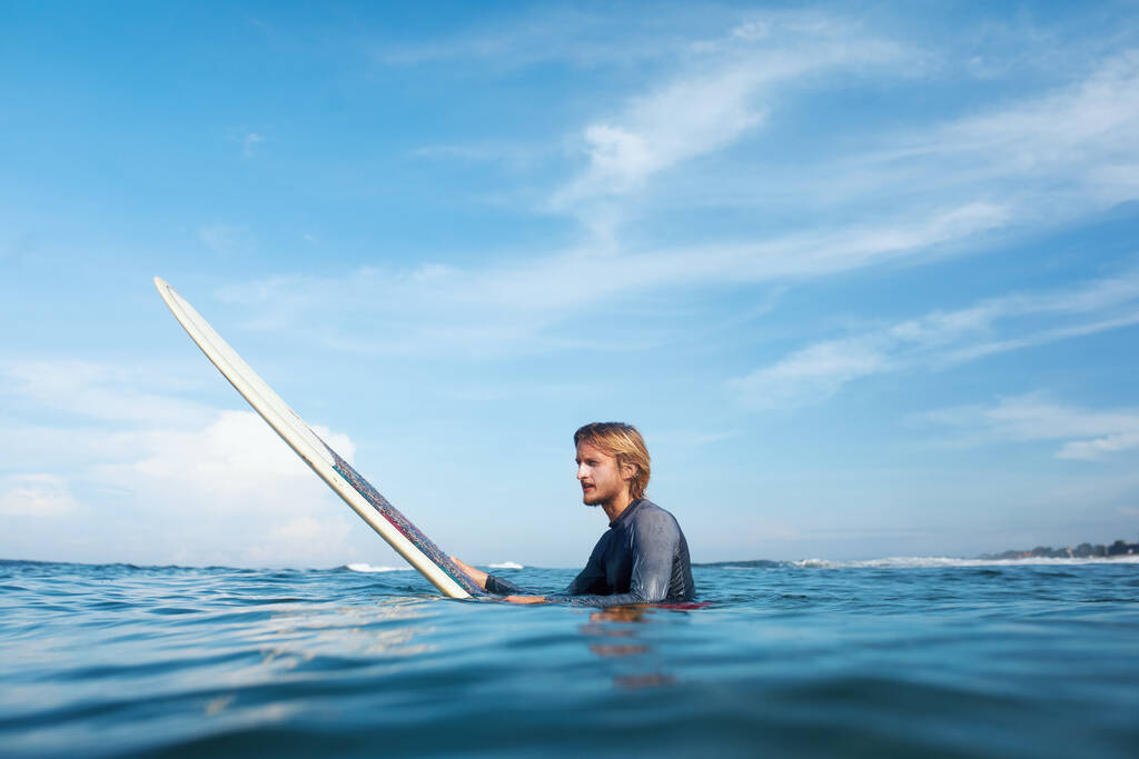 Surfer. Jongeman in wetsuit op wit surfbordportret. Knappe vent in de oceaan. Blauwe zee en lucht met zachte wolken op de achtergrond. - Foto, afbeelding