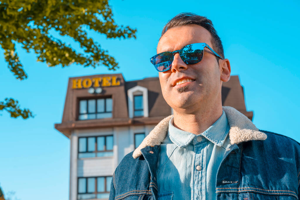 Lifestyle, een jonge blanke man in een jasje en zonnebril met een hotel op de achtergrond - Foto, afbeelding