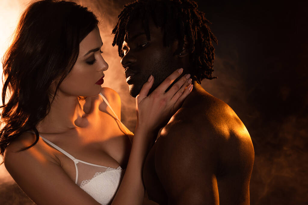 hermosa pareja seductora multicultural abrazándose en la cama en la oscuridad con humo
  - Foto, Imagen