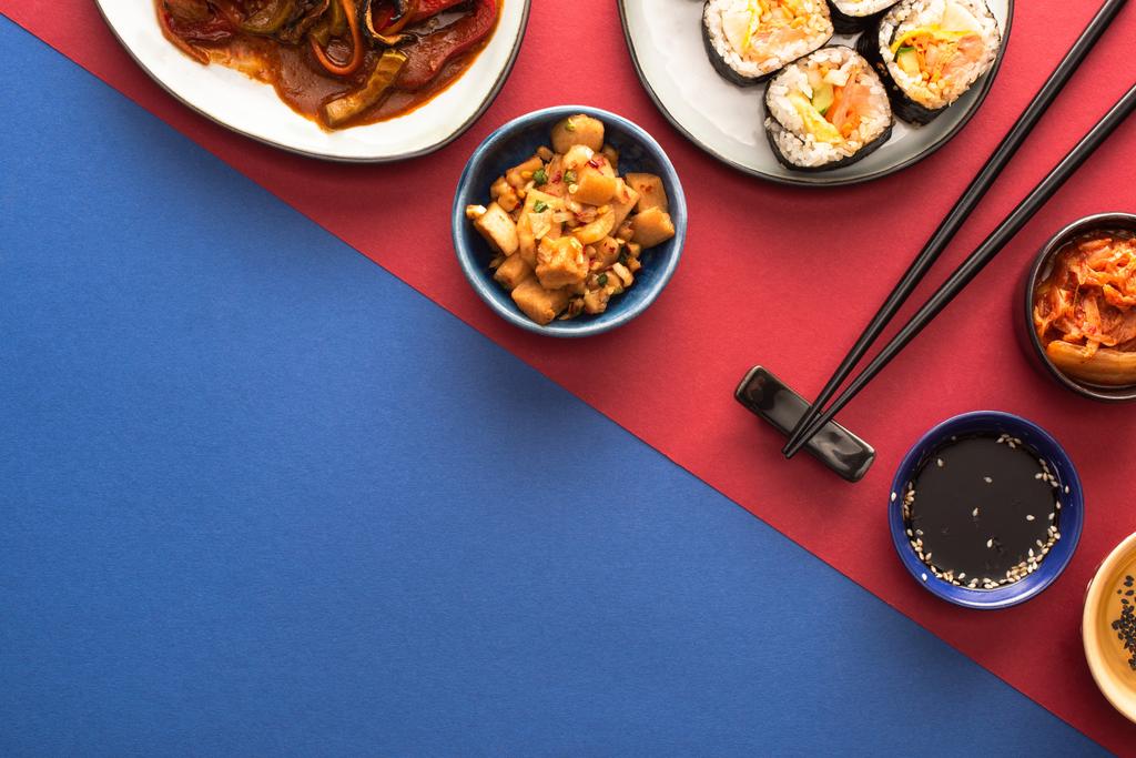 Draufsicht auf Schalen und Teller mit koreanischem Kimchi und Kimbap auf blau und purpurrot   - Foto, Bild
