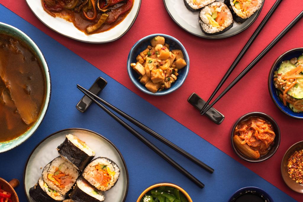 vrchní pohled na hůlky v blízkosti ramen, kimbap a korejské vedlejší pokrmy na modré a karmínové  - Fotografie, Obrázek