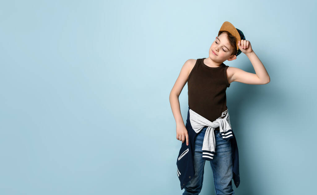 Active kid dressed in sporty style stands looking aside and touching cap on his head, creating prank or mischief. Studio shot isolated on blue - Photo, Image