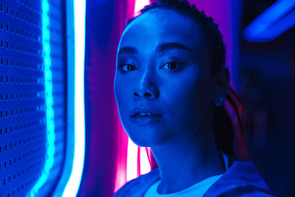 Gyönyörű, fiatal ázsiai nő, hosszú, sötét hajú, infravörös neon fal előtt áll, a kamerába néz. - Fotó, kép