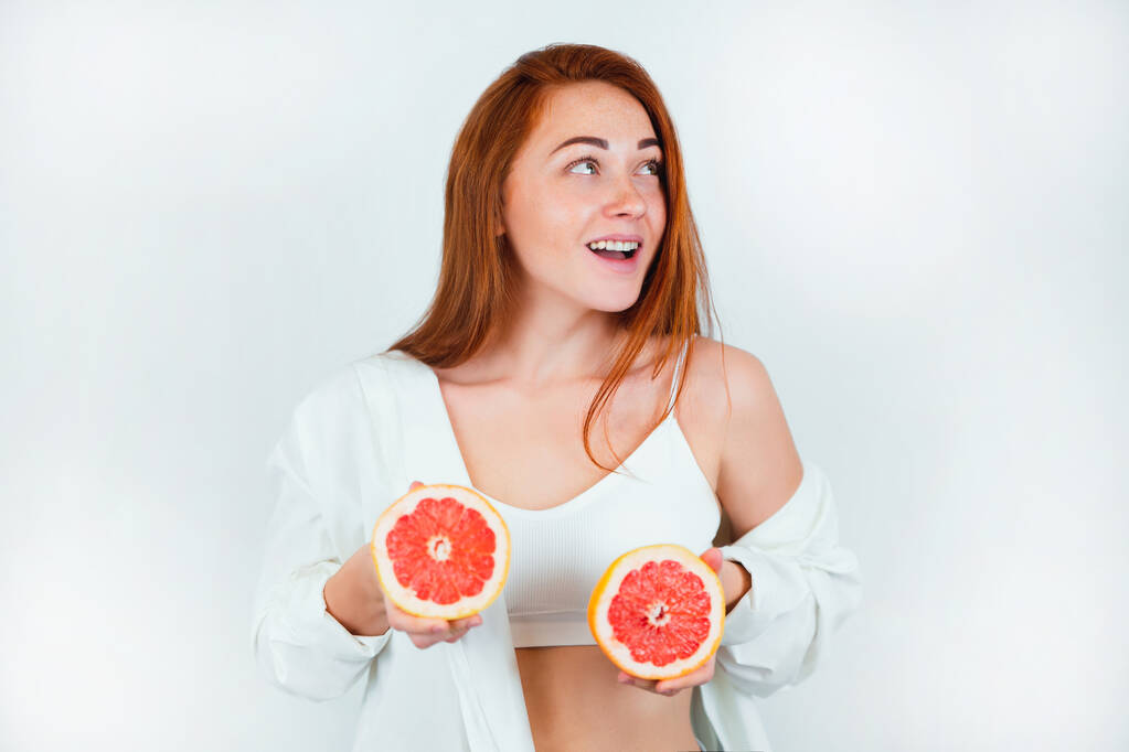 portrét rusovlasé mladé rozesmáté ženy, která vypadá přirozeně, stojící na osamělém bílém podkladu, držící v rukou půlky grapefruitu v blízkosti obličeje, koncept portrétu krásy. - Fotografie, Obrázek