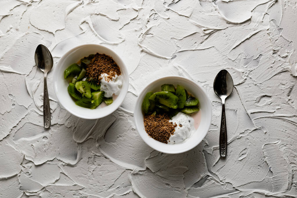 A pair of small dessert bowls with fresh kiwi fruit, white ice cream, yogurt and chocolate chips on a white stone background with teaspoons - Photo, Image