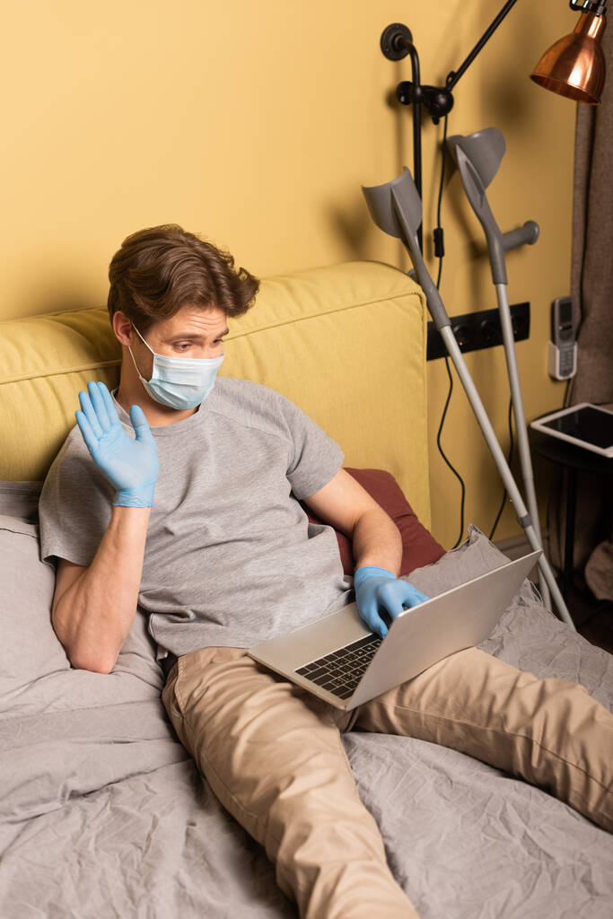 Tıbbi maskeli engelli bir adamın yatak odasındaki koltuk değneklerinin yanında dizüstü bilgisayarında görüntüsü var. - Fotoğraf, Görsel