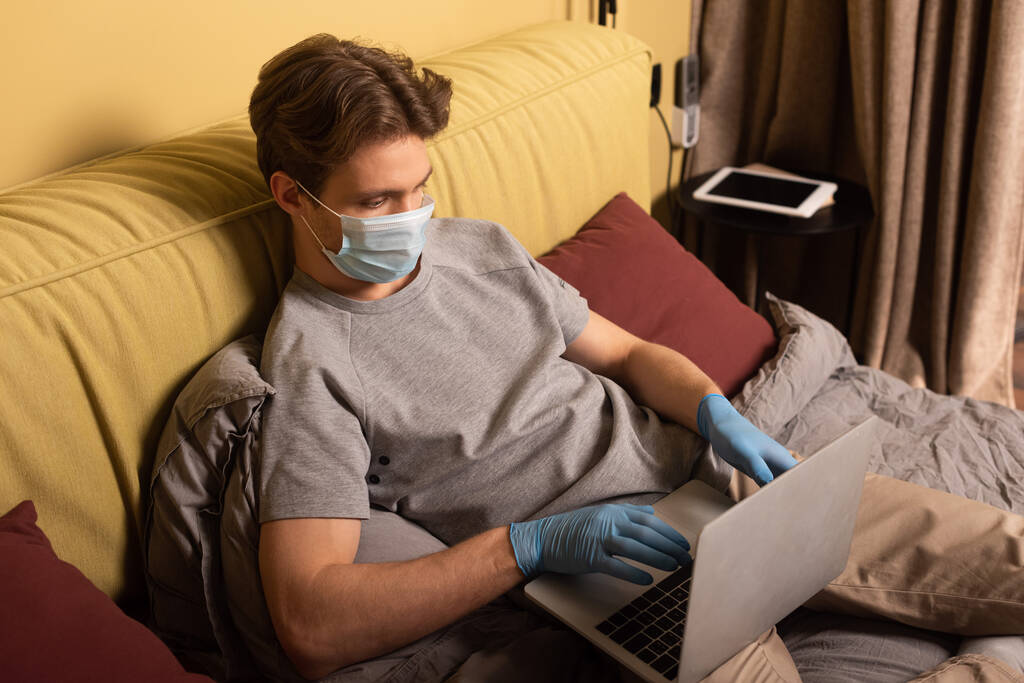 医療用マスクとラテックス手袋の男はベッドの上でノートパソコンで作業  - 写真・画像