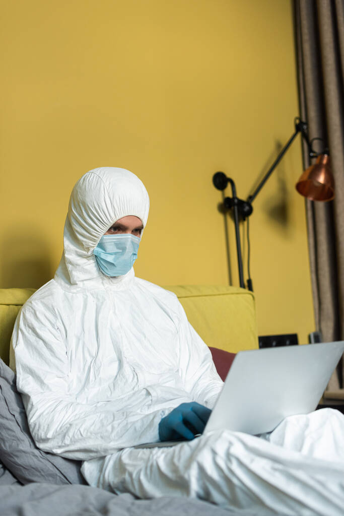 Enfoque selectivo del hombre en traje de materiales peligrosos y máscara médica que trabaja en el ordenador portátil en la cama
  - Foto, Imagen