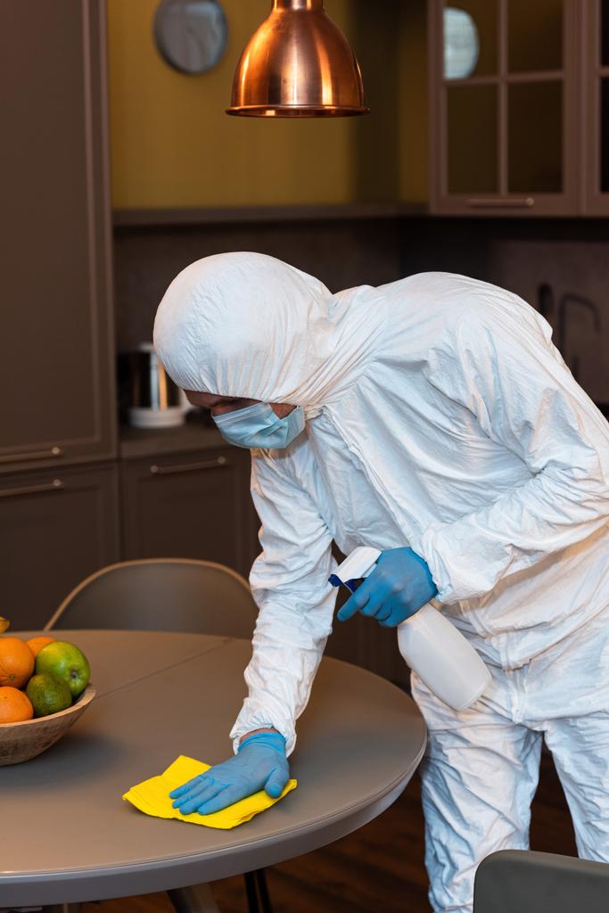 Άνδρας με στολή, ιατρική μάσκα και γάντια λάτεξ καθαρίζοντας τραπέζι με κουρέλι και απορρυπαντικό στην κουζίνα  - Φωτογραφία, εικόνα