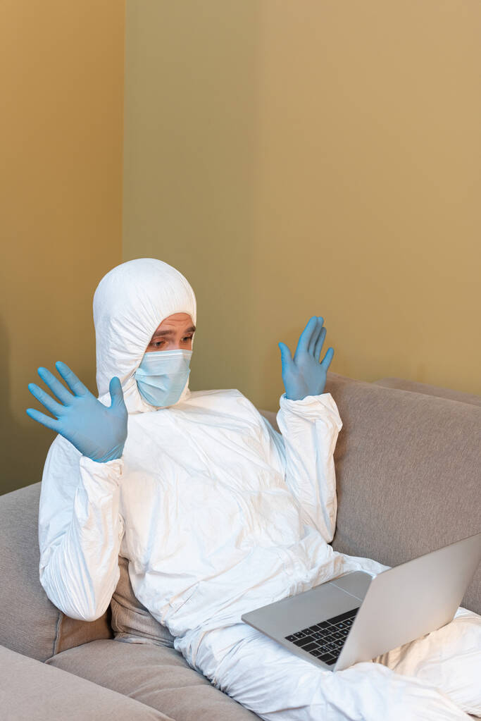 Man in hazmat pak en medisch masker zwaaiende handen tijdens videogesprek op laptop  - Foto, afbeelding