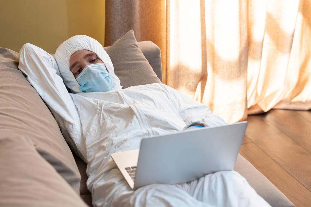 Селективный фокус человека в защитном костюме и медицинской маске с помощью ноутбука на диване
  - Фото, изображение