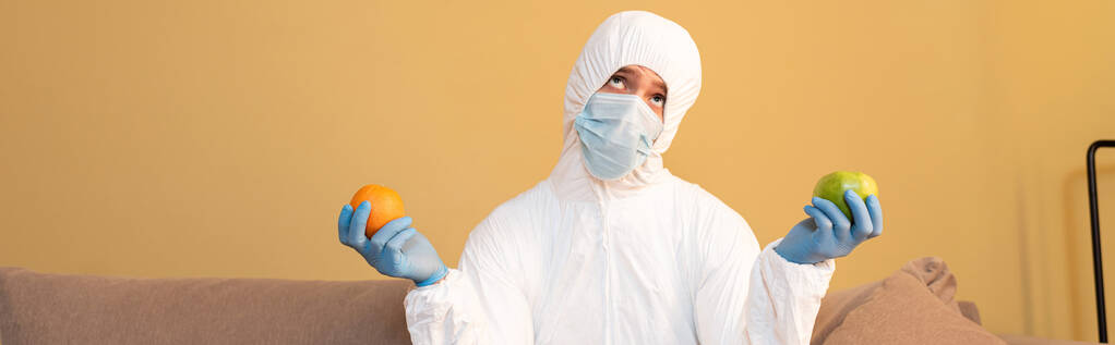 Πανοραμικός προσανατολισμός στοχαστικού άνδρα με στολή και ιατρική μάσκα που κρατάει μήλο και πορτοκάλι στο σπίτι  - Φωτογραφία, εικόνα