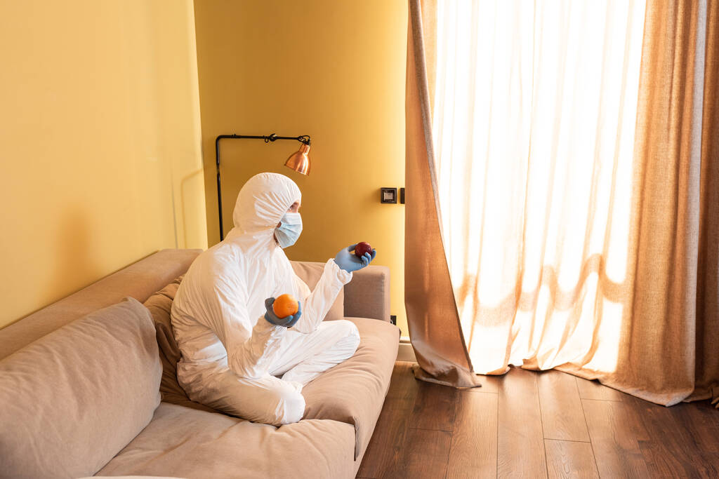 Hombre pensativo con máscara médica, guantes de látex y traje de hazmat sosteniendo manzana y naranja mientras está sentado en el sofá
  - Foto, Imagen