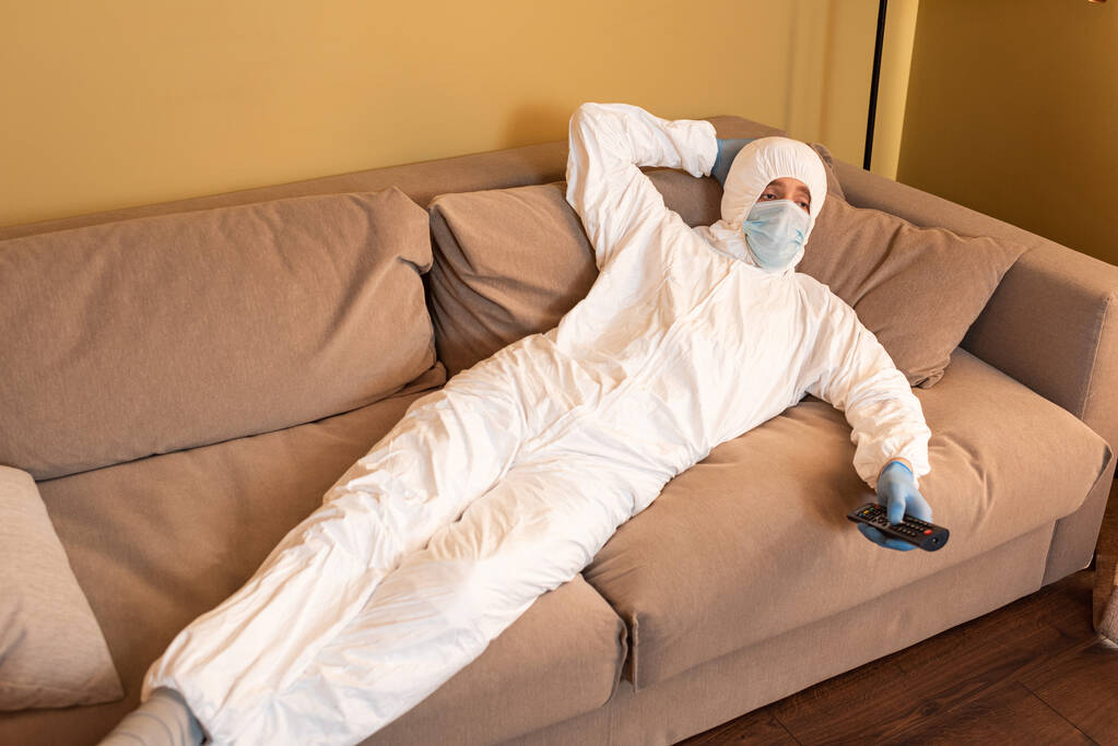 Tıbbi maskeli bir adam, koruyucu giysi ve lateks eldivenler. Evdeki kanepede televizyon seyrediyor.  - Fotoğraf, Görsel