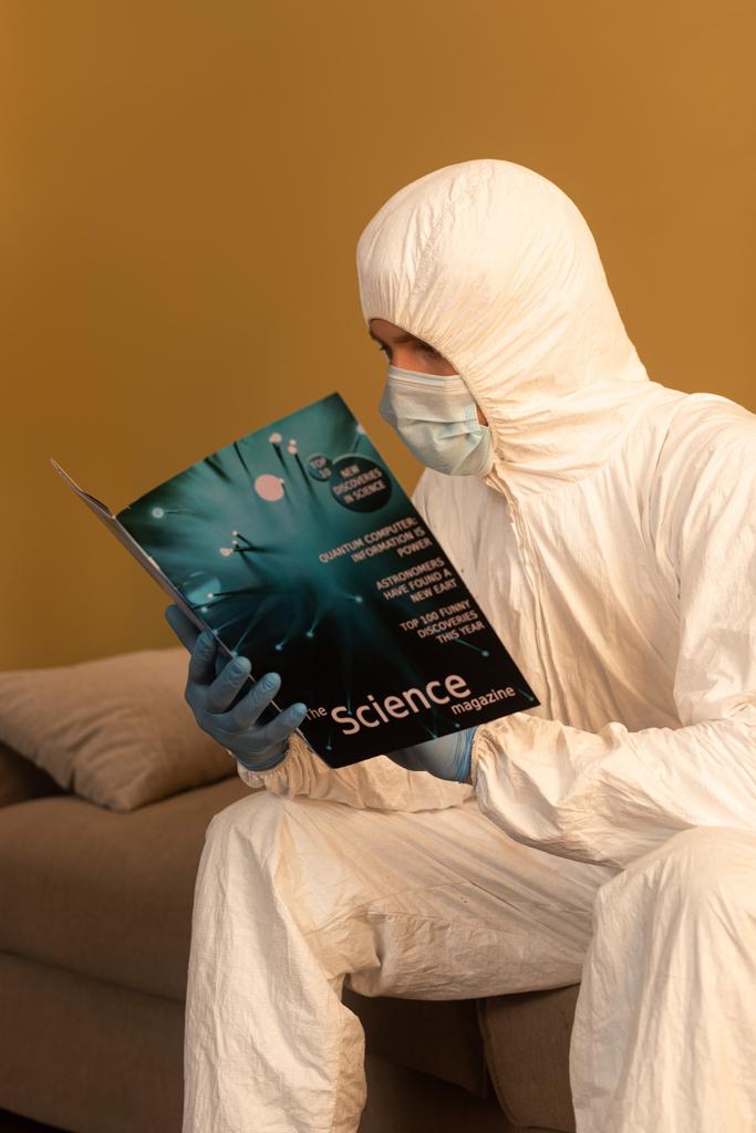 Homem em máscara médica e terno hazmat leitura revista científica no sofá em casa
  - Foto, Imagem
