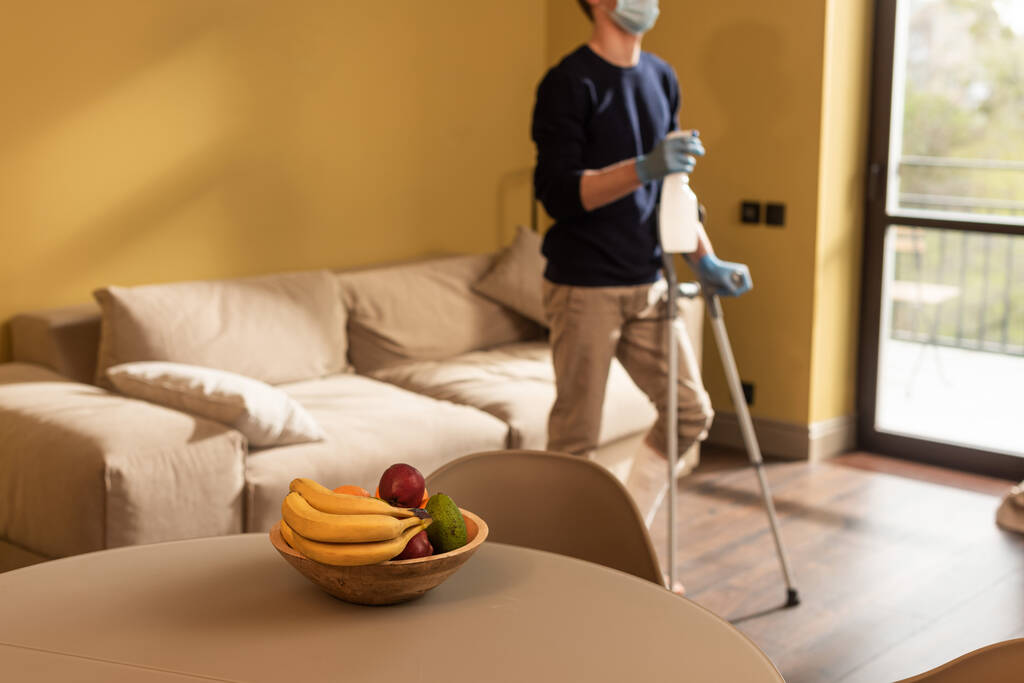 Selective focus of disabled man in medical mask holding bottle of detergent and crutches in living room - Photo, Image