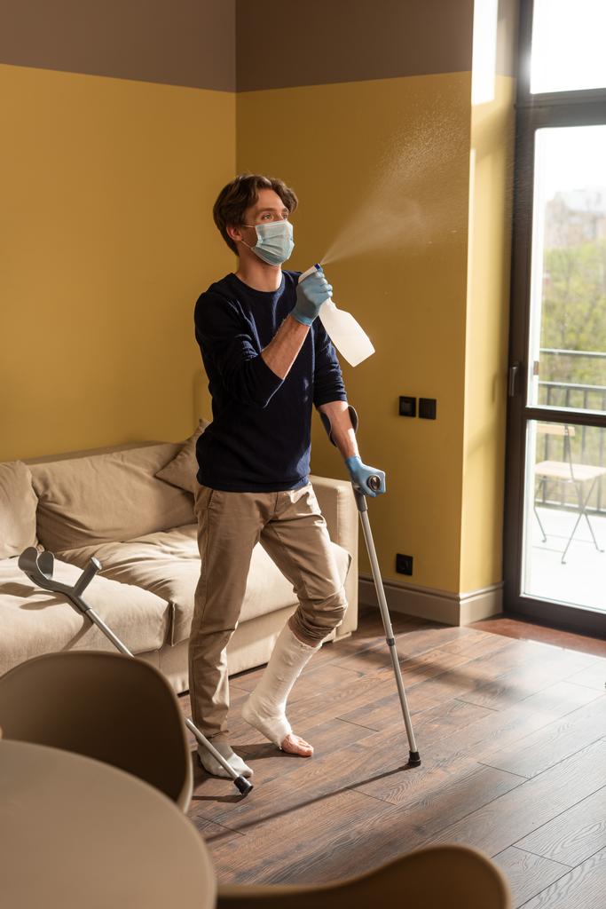 Selective focus of disabled man in medical mask holding crutches and spraying detergent at home  - Photo, Image