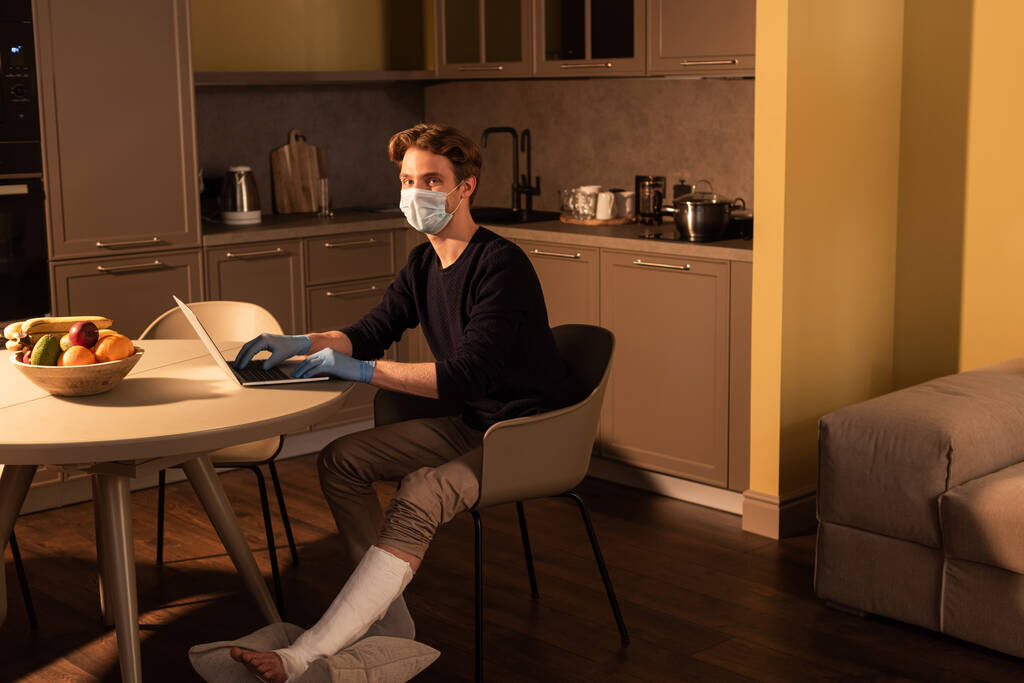 Tıbbi maskeli ve alçı bandajlı bir adam mutfaktaki dizüstü bilgisayarı kullanıyor.  - Fotoğraf, Görsel
